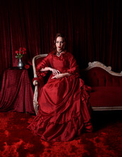   Portrait Of Pretty Female Model With Red Hair Wearing Glamorous Historical Victorian Red Ballgown.  Posing With A Moody Dark Background.