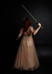 Wall Mural -  Full length portrait of pretty female model with red hair wearing glamorous fantasy tulle gown and crown.  Posing with a moody dark background.