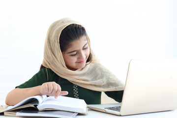 Adorable smiling Pakistani Muslim girl with beautiful eyes wearing hijab, studying online, doing homework by using laptop computer, student kid reading book on white background.