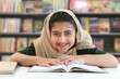 Leinwandbild Motiv Adorable smiling Pakistani Muslim girl with beautiful eyes wearing hijab, studying and doing homework on table, happy student kid reading book on blurred background of bookcase in library.