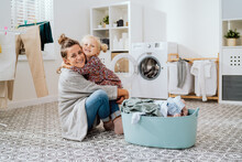 Mother Sits On Laundry Room Floor With Little Daughter, A Cute Little Girl With Blonde Hair Tied Up In A Bun Grabs The Woman By Neck Hugs Her Tightly They Spend Time Doing Housework Sorting Clothes.
