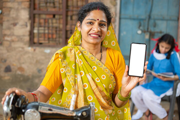 Portrait of happy traditional indian woman wearing sari using sewing machine while showing smart phone with empty display to put advertisement, female holding blank android phone screen.