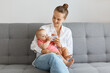 © sementsova321 - Indoor shot of smiling delighted woman with bun hairstyle wearing white shirt and jeans, holding her toddler daughter and giving to her bottle in bottle.