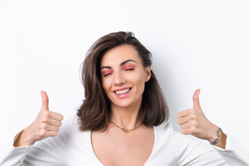 Wall Mural - Young woman in a cocktail dress, gold chain and bright spring pink makeup on a white background. Cheerfully smiling showing thumbs up