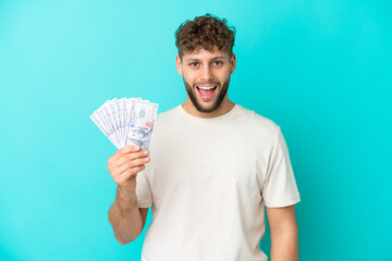 Poster - Young caucasian man taking a lot of money isolated on blue background with surprise facial expression