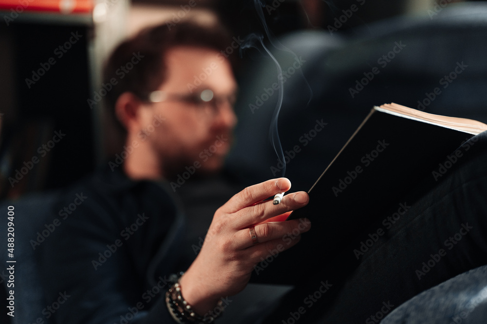 Man smoking and reading, holding a book and a cigarette in hands while  lying on a couch. Focus on cigarette. Stock Photo
