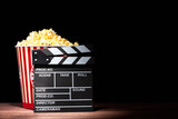 Fototapeta Na drzwi - Cinema concept. Popcorn in a box and movie clapper on wooden table under beam of light against black background