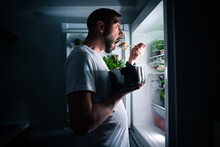 Hungry Man Eating Food At Night From Open Fridge. Man Taking Midnight Snack From Refrigerator