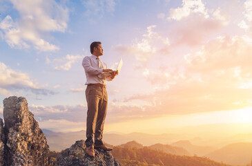 Businessman working outdoors with laptop computer standing on top rock mountain at sunset background. Work and vacation.