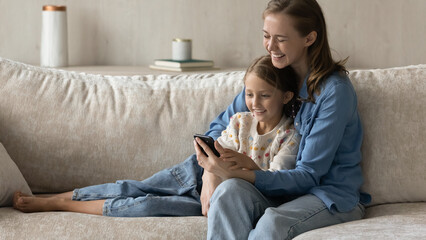 Wall Mural - Happy laughing mom and smiling kid using online ecommerce app on smartphone, shopping on internet together, watching funny video, media content on mobile phone, relaxing on sofa at home