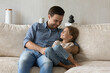 © fizkes - Cheerful daddy tickling and cuddling happy little daughter kid on comfortable couch, hugging girl with tenderness, love, care, talking to child, having fun. Fatherhood, family leisure time concept