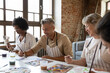 © fizkes - Happy handsome man in apron drawing picture with paints on paper, enjoying creative art master class workshop with smiling multiracial young and old people, sitting together at table in loft studio.