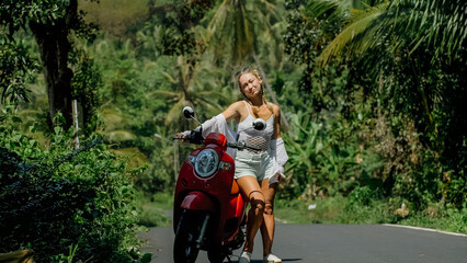 Wall Mural - Woman on red scooter in white clothes drive on forest road trail. Dancing trip. One girl caucasian tourist in sunglasses dance, relax, rest near motorbike. Asia Thailand ride tourism. Motorcycle rent.