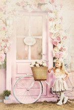 A Blonde Preschool Girl Stands At A Vintage Door With A Bicycle. Floral Decor. Design In The Form Of A Postcard.