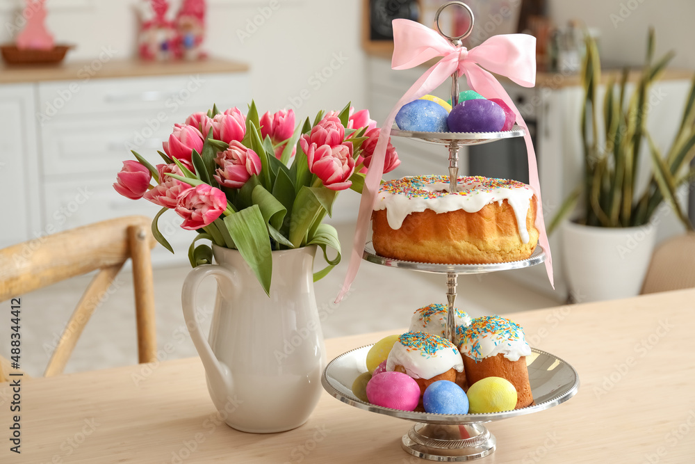 Stand with Easter cakes, eggs and tulips on dining table in kitchen