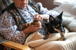 A senior woman with a little dog on her lap sits by the window and knits a warm sock
