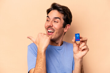 Wall Mural - Young caucasian man holding a batterie isolated on beige background points with thumb finger away, laughing and carefree.