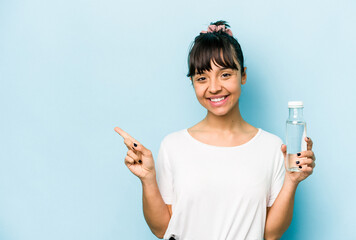 Young hispanic woman holding a bottle of water isolated on blue background smiling and pointing aside, showing something at blank space.