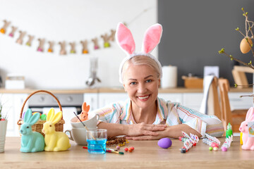 Wall Mural - Mature woman with bunny ears, Easter eggs and paints at home