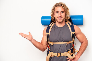 Wall Mural - Young mountaineer caucasian man with a big backpack isolated on white background showing a copy space on a palm and holding another hand on waist.