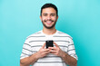 © luismolinero - Young Brazilian man isolated on blue background looking at the camera and smiling while using the mobile