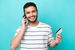 © luismolinero - Young Brazilian man isolated on blue background keeping a conversation with the mobile phone and holding a credit card