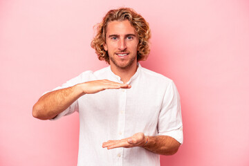 Wall Mural - Young caucasian man isolated on pink background holding something with both hands, product presentation.