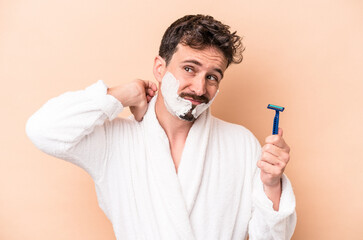 Wall Mural - Young caucasian man wearing shaving foam and holding razor blade isolated on beige background touching back of head, thinking and making a choice.