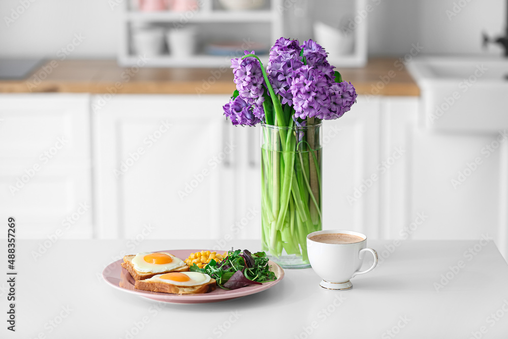 Vase with flowers and delicious breakfast on table in kitchen