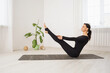Woman in black sports overalls practicing yoga doing Paripurna Navasana exercise, boat pose, exercising while sitting on a mat in the room