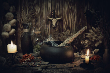 Authentic forest witch altar. Burning candles, dry herbs, iron cauldron, smoking mugwort, bulls skull mojo, bunch of garlic in wooden background in the dark, low key, selective focus.