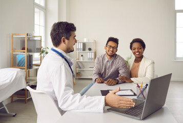 Wall Mural - Smiling male doctor in medical uniform show checkup results on laptop screen to ethnic couple patients in clinic. Man GP or therapist consult spouses in modern hospital. Healthcare and medicine.