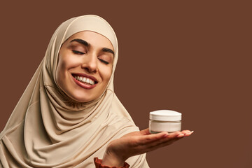 Portrait of beautiful modern muslim woman with natural make-up dressed in beige hijab smiling with closed eyes and holding cream in hand on brown background. Facial skin care, female beauty.