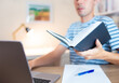 © Marcio - Student sitting at desk in his bedroom and using laptop to study and holding a book in hands . Studying or preparing for the test at home.