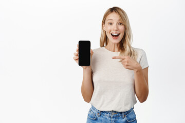Wall Mural - Enthusiastic smiling woman showing mobile phone screen, smartphone app interface, concept of technology and cellular, standing over white background