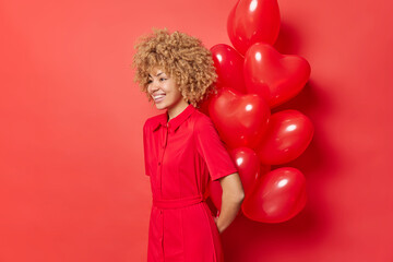 Wall Mural - Horizontal shot of cheerful curly haired woman prepares surprise for lover on Valentines Day holds bunch of heart balloons behind back smiles happily wears dress isolated over vivid red background