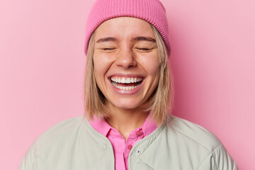 Wall Mural - Positive authentic European woman smiles broadly shows white teeth giggles happily with closed eyes wears hat and jacket poses in studio against pink background. People and emotions concept.