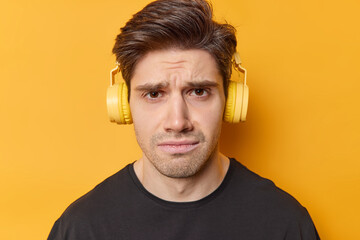 Studio shot of discontent adult man with dark hair wears casual black t shirt listens music via headphones isolated over yellow bbackground expresses negative emotions. People and hobby concept