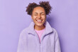 Leinwandbild Motiv Cute positive young woman with hair buns smiles broadly keeps eyes closed feels very happy dressed in winter coat isolated over purple background being in good mood. People and good mood concept