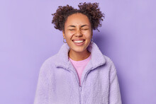 Cute Positive Young Woman With Hair Buns Smiles Broadly Keeps Eyes Closed Feels Very Happy Dressed In Winter Coat Isolated Over Purple Background Being In Good Mood. People And Good Mood Concept
