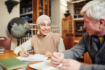 Portrait of smiling Muslim woman wearing head covering while studying in college library with senior professor