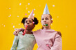 Portrait of two girls wearing party hats while celebrating Birthday against pop yellow background in studio