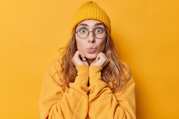 Poster - Portrait of surprised young woman keeps hands under chin has lips folded looks shocked wears hat and sweatshirt big round spectacles isolated over yellow background. Human reactions concept.