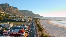 Low Altitude Aerial Shot Flying Directly Overhead The Busy Main Street Of Camps Bay's Beachfront In Cape Town During Sunset With The Twelve Apostles As A Backdrop.