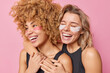 Horizontal shot of cheerful young women embrace and have fun apply beauty hydrogel patches under eyes take care of delicate skin smile broadly wear black t shirts isolated over pink background