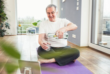 Happy Senior Man Sitting On Exercise Mat Pouring Water In Drinking Glass At Home