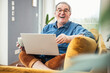 © Westend61 - Cheerful senior man with laptop and coffee mug sitting on sofa in living room