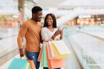 Wall Mural - Portrait of excited black spouses using phone holding shopping bags