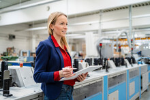Smiling Blond Businesswoman With Tablet PC Standing By Machinery In Factory