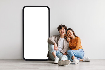 Loving asian couple sitting near big cellphone with empty white screen, embracing, recommending app or cool website